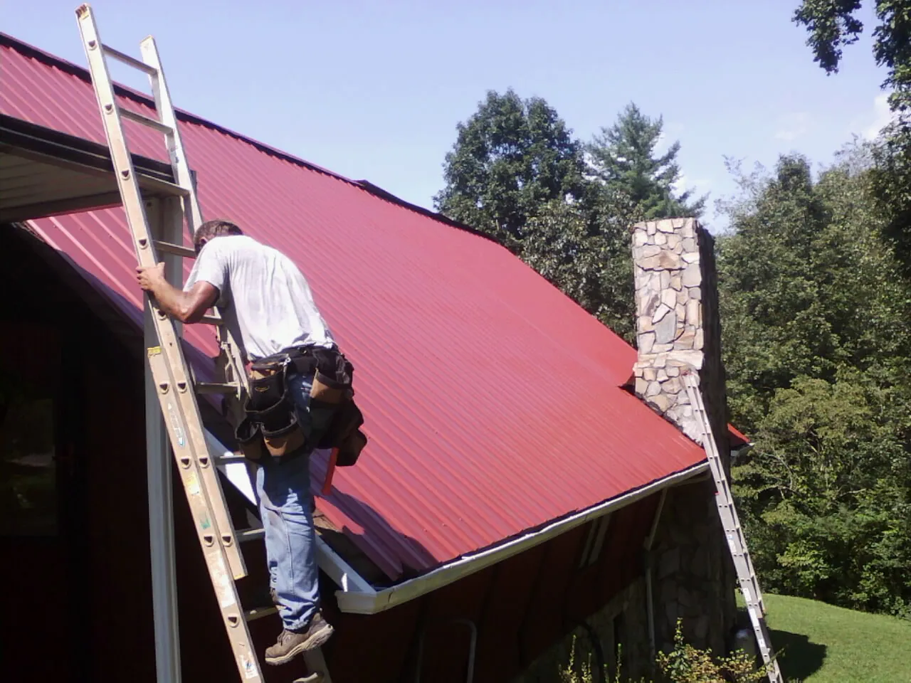 Roofer on a ladder beside a red metal roof and stone chimney — Soffit & Fascia Repair by Roofing Brothers in Carlock